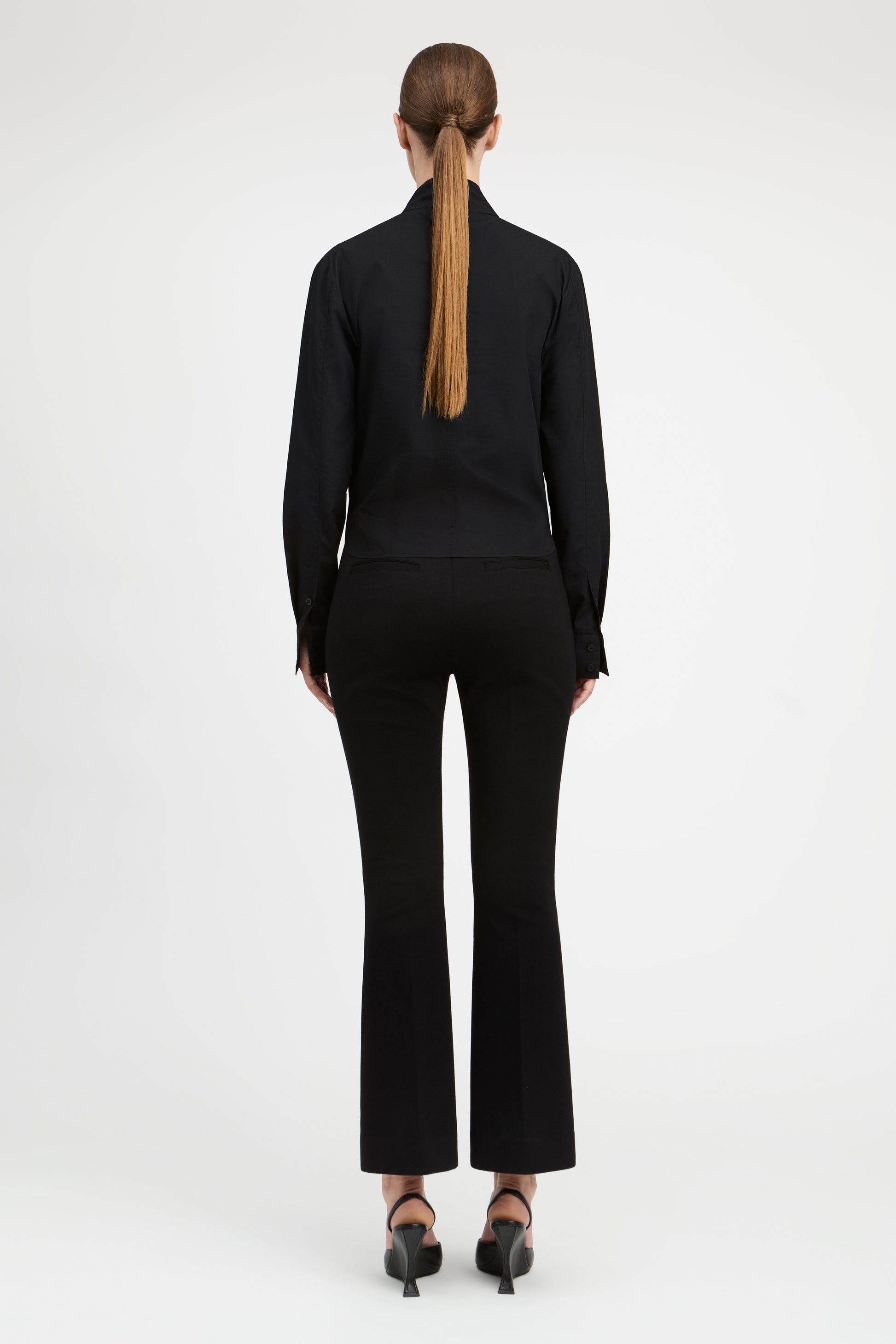 A woman viewed from the back wearing a black long-sleeve mock-neck top and matching black flared pants, paired with black heels, against a plain light gray studio background.