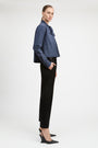 A model is shown in profile wearing a blue and white vertically striped button-down shirt with a bow collar, paired with black trousers and black wedge heels, against a plain light gray studio background.