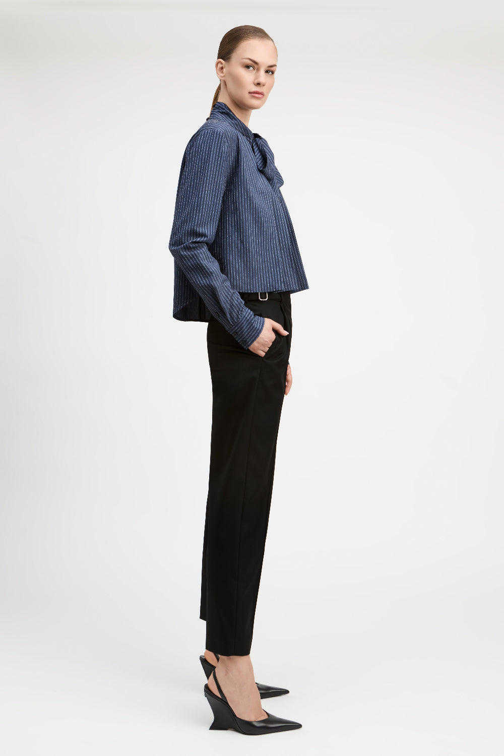 A model is shown in profile wearing a blue and white vertically striped button-down shirt with a bow collar, paired with black trousers and black wedge heels, against a plain light gray studio background.