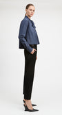 A model is shown in profile wearing a blue and white vertically striped button-down shirt with a bow collar, paired with black trousers and black wedge heels, against a plain light gray studio background.