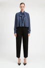 A model stands against a light gray studio background wearing a blue and white vertically striped long-sleeve blouse with a large bow tie-neck detail, paired with black cropped trousers and black pointed heels.