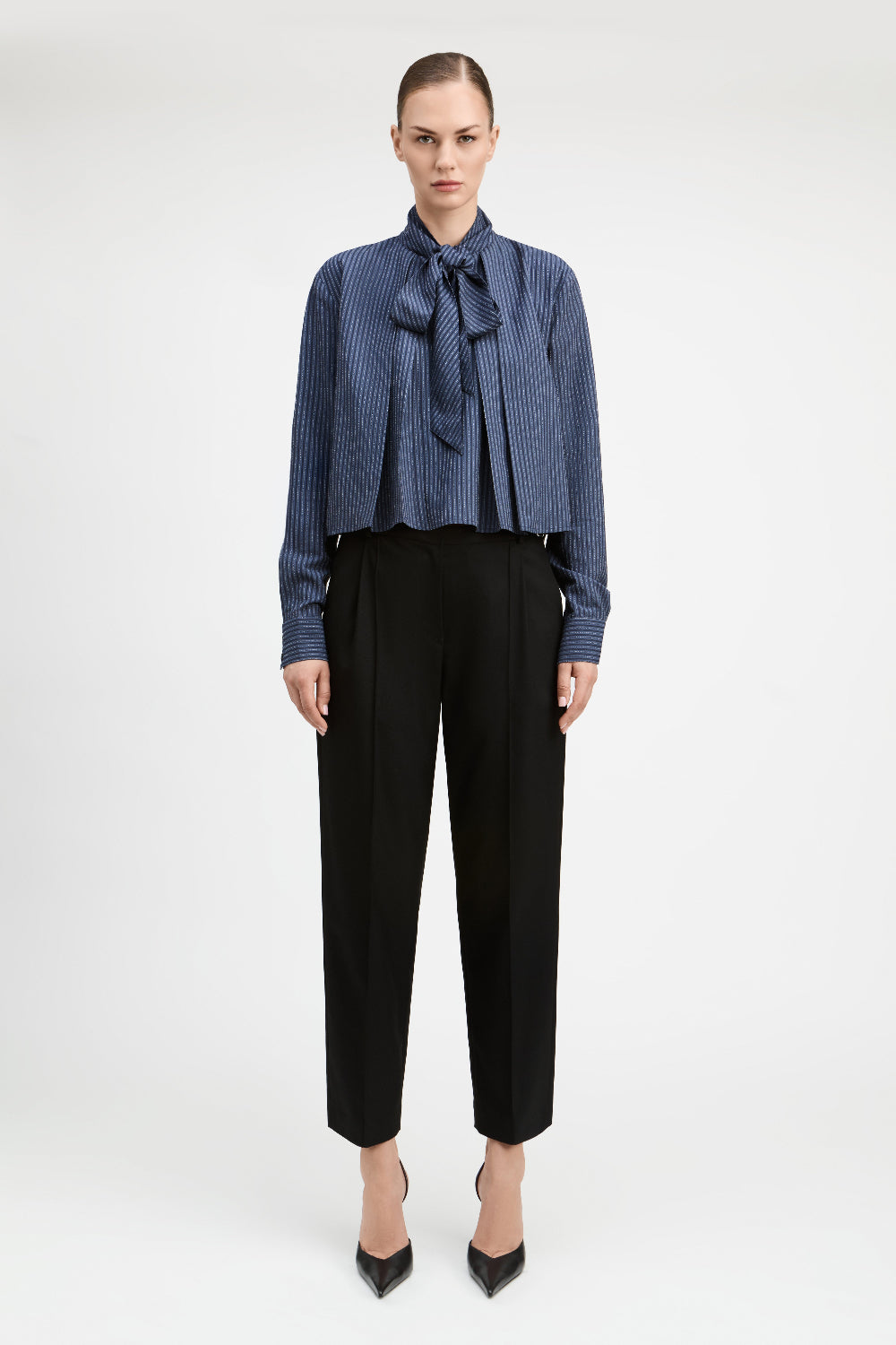 A model stands against a light gray studio background wearing a blue and white vertically striped long-sleeve blouse with a large bow tie-neck detail, paired with black cropped trousers and black pointed heels.