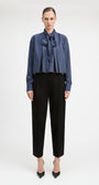 A model stands against a light gray studio background wearing a blue and white vertically striped long-sleeve blouse with a large bow tie-neck detail, paired with black cropped trousers and black pointed heels.