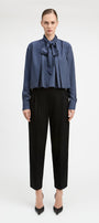 A model stands against a light gray studio background wearing a blue and white vertically striped long-sleeve blouse with a large bow tie-neck detail, paired with black cropped trousers and black pointed heels.
