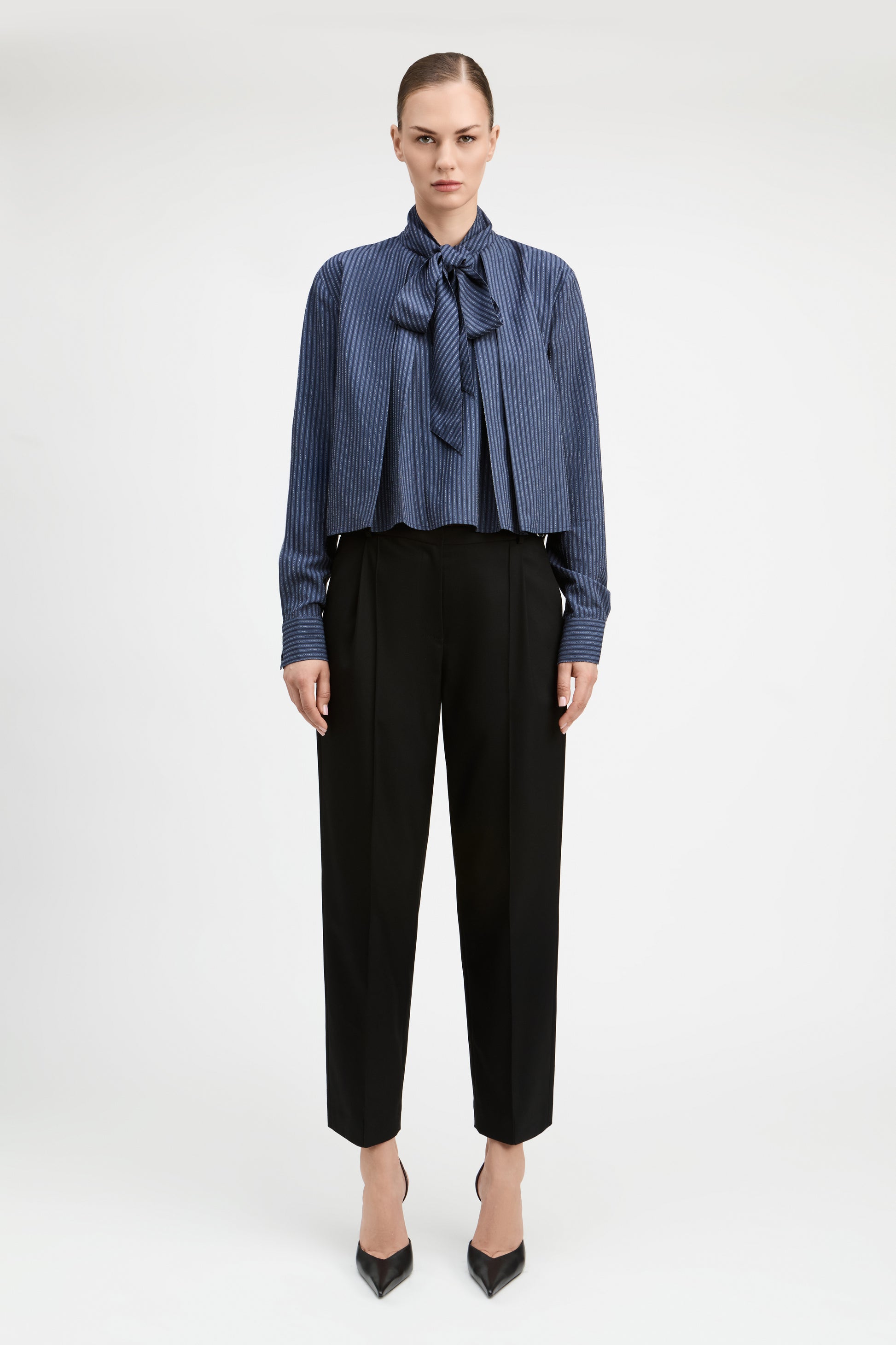 A model stands against a light gray studio background wearing a blue and white vertically striped long-sleeve blouse with a large bow tie-neck detail, paired with black cropped trousers and black pointed heels.