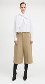A model stands against a light gray studio background wearing a white long-sleeve blouse with a large bow at the collar, beige wide-leg cropped pants, and black heeled boots.