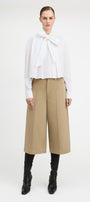 A model stands against a light gray studio background wearing a white long-sleeve blouse with a large bow at the collar, beige wide-leg cropped pants, and black heeled boots.