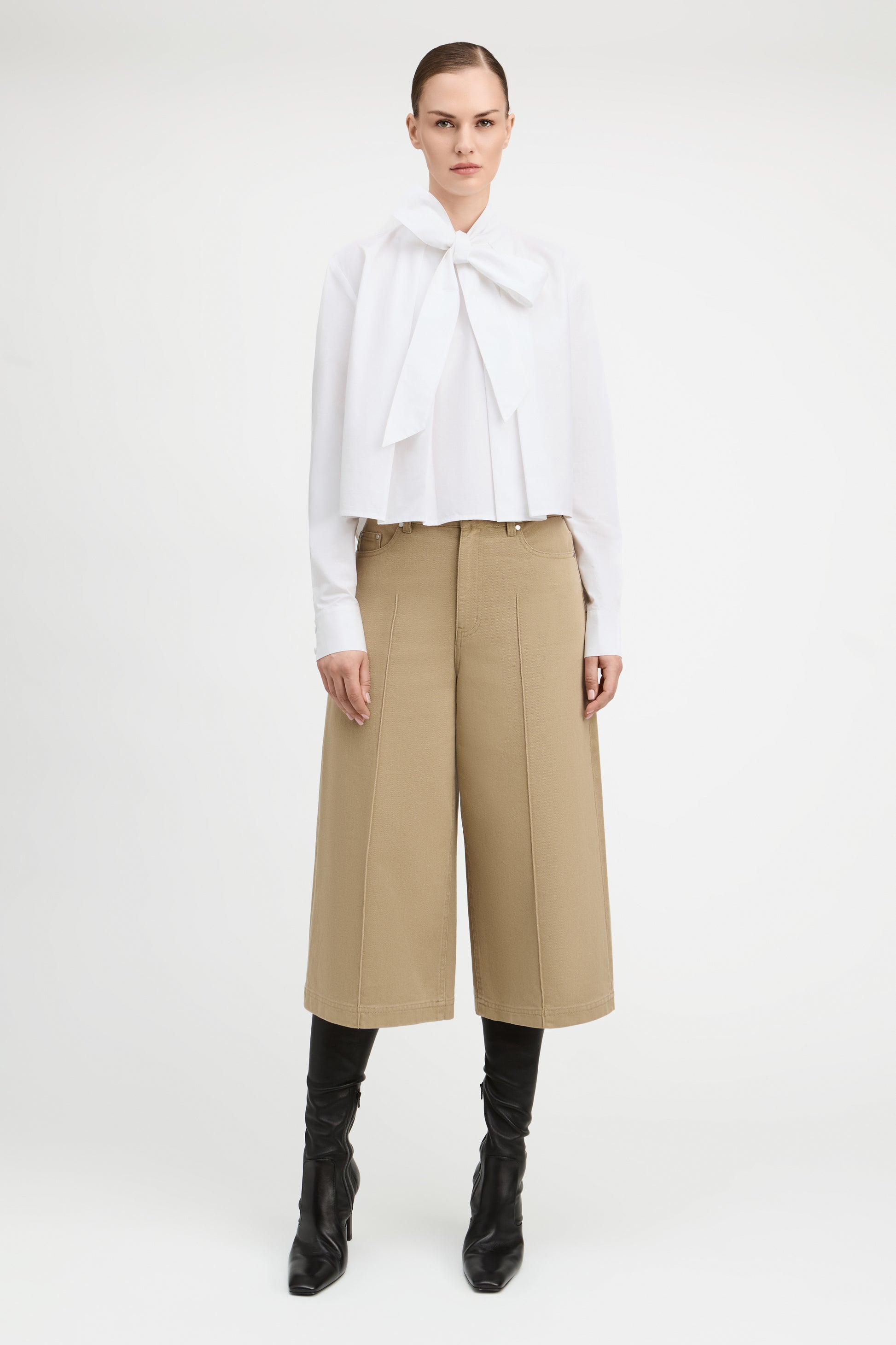 A model stands against a light gray studio background wearing a white long-sleeve blouse with a large bow at the collar, beige wide-leg cropped pants, and black heeled boots.