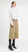 A model is shown in profile wearing a white long-sleeve blouse with a large bow at the collar, paired with a beige wide-leg midi skirt and black knee-high heeled boots, against a plain white studio background.