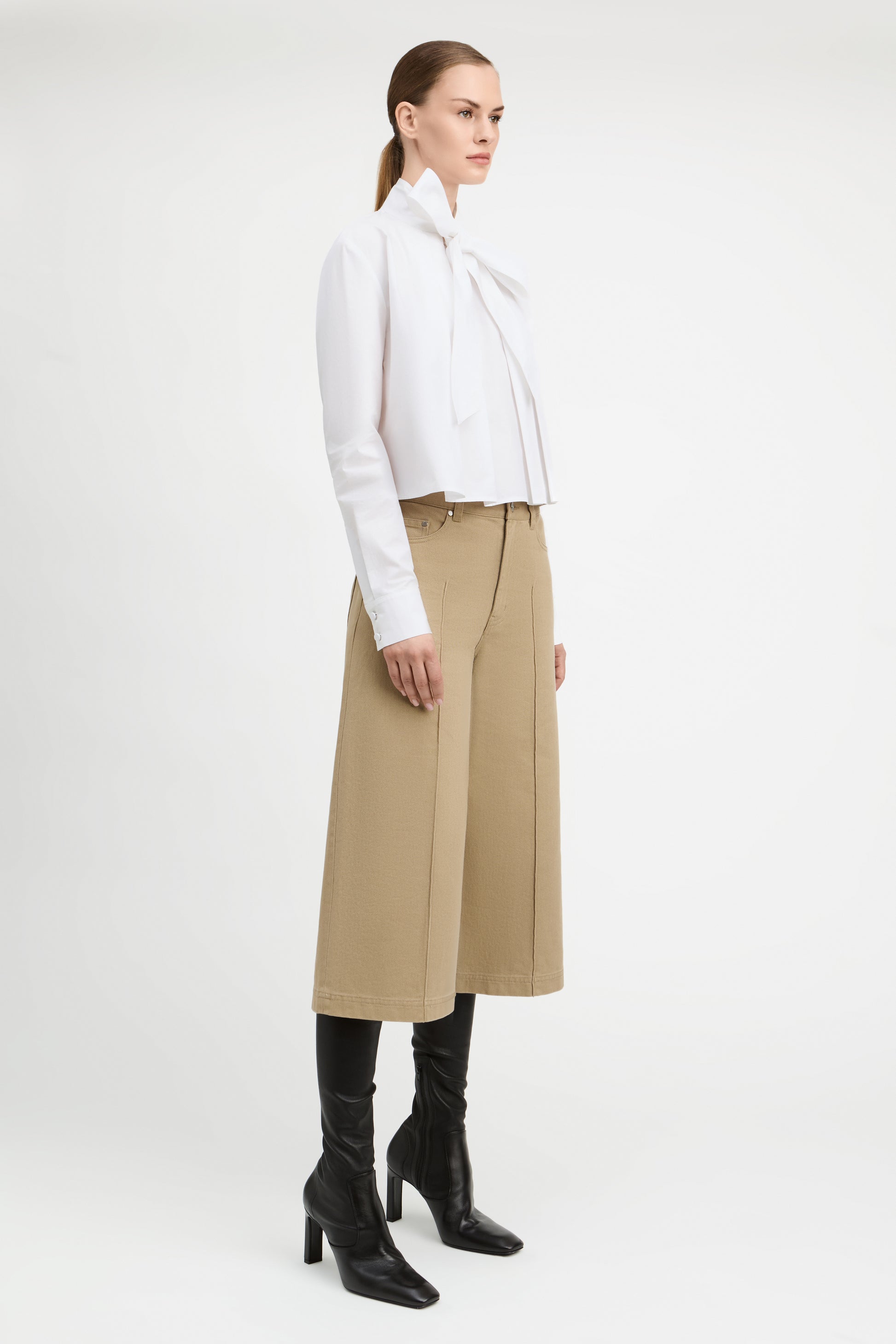 A model is shown in profile wearing a white long-sleeve blouse with a large bow at the collar, paired with a beige wide-leg midi skirt and black knee-high heeled boots, against a plain white studio background.