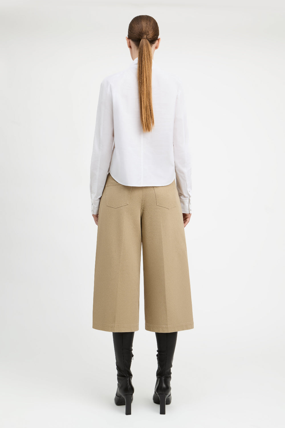 A woman shown from the back wearing a white long-sleeve blouse, beige wide-leg cropped pants, and black heeled boots against a plain light gray studio background.