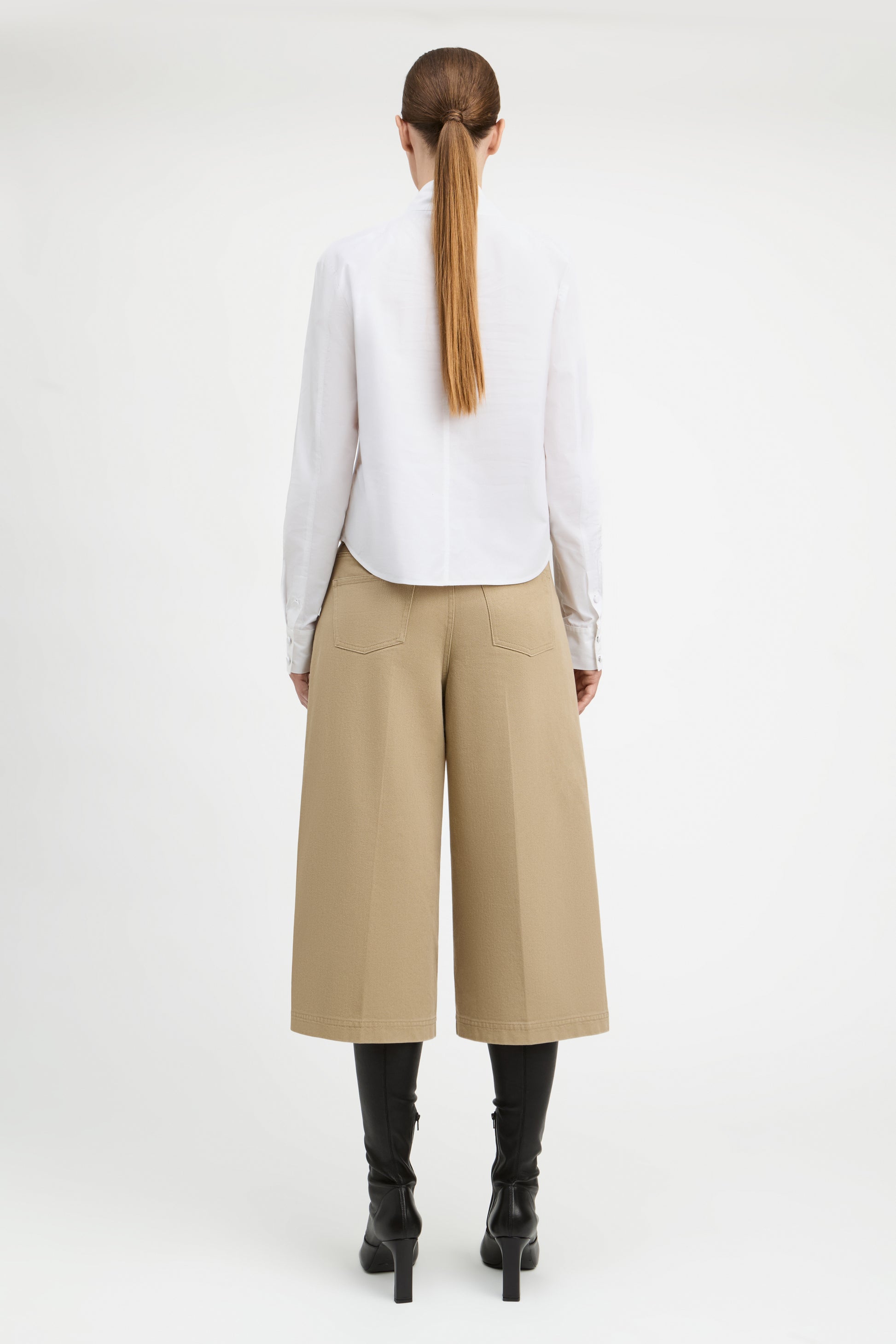 A woman shown from the back wearing a white long-sleeve blouse, beige wide-leg cropped pants, and black heeled boots against a plain light gray studio background.