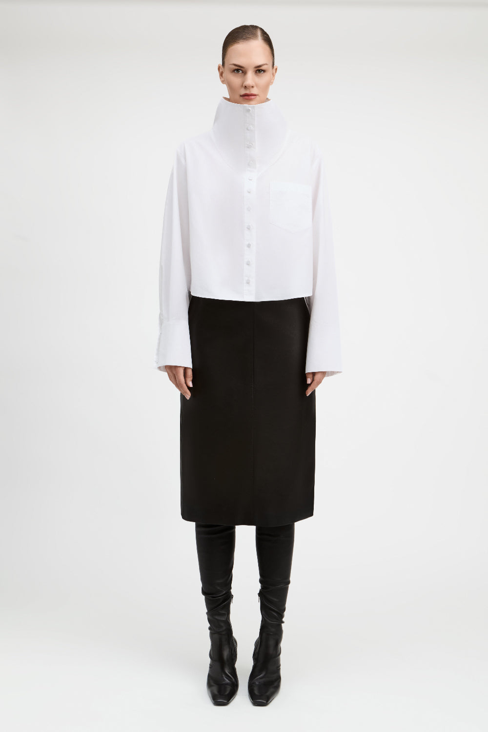 A model stands against a plain light gray studio background wearing a white long-sleeve high-neck button-front top with a chest pocket, paired with a black midi skirt, black leggings, and black pointed boots.