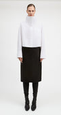 A model stands against a plain light gray studio background wearing a white long-sleeve high-neck button-front top with a chest pocket, paired with a black midi skirt, black leggings, and black pointed boots.
