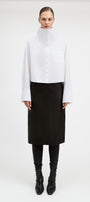 A model stands against a plain light gray studio background wearing a white long-sleeve high-neck button-front top with a chest pocket, paired with a black midi skirt, black leggings, and black pointed boots.