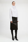 A model stands against a light gray studio background wearing a white asymmetrical mock-neck blouse with long sleeves, a black midi skirt, black leggings, and black heeled ankle boots.