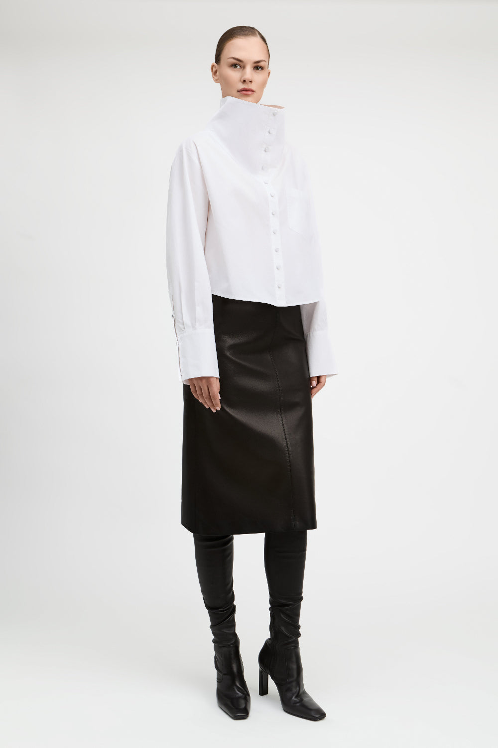 A model stands against a light gray studio background wearing a white asymmetrical mock-neck blouse with long sleeves, a black midi skirt, black leggings, and black heeled ankle boots.
