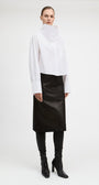A model stands against a light gray studio background wearing a white asymmetrical mock-neck blouse with long sleeves, a black midi skirt, black leggings, and black heeled ankle boots.