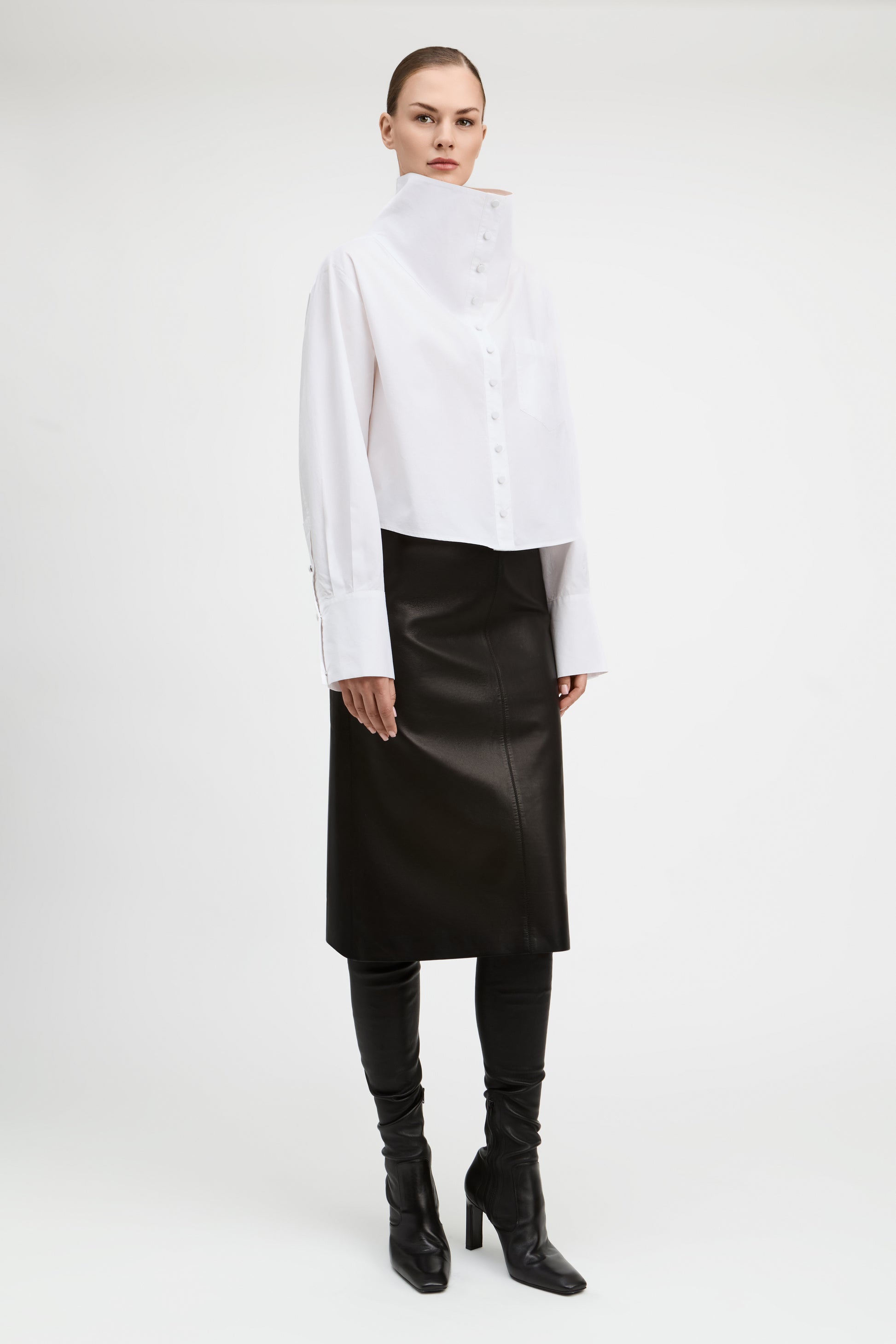 A model stands against a light gray studio background wearing a white asymmetrical mock-neck blouse with long sleeves, a black midi skirt, black leggings, and black heeled ankle boots.