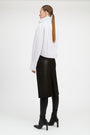 A woman shown from the back wearing a white high-neck long-sleeve top, a black midi skirt, black leggings, and black heeled ankle boots, photographed against a plain light gray studio background.