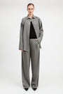 A model stands against a light gray studio background wearing a matching gray two-piece set consisting of a long-sleeve button-front overshirt and wide-leg trousers, styled over a black top and black pointed shoes.