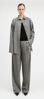 A model stands against a light gray studio background wearing a matching gray two-piece set consisting of a long-sleeve button-front overshirt and wide-leg trousers, styled over a black top and black pointed shoes.