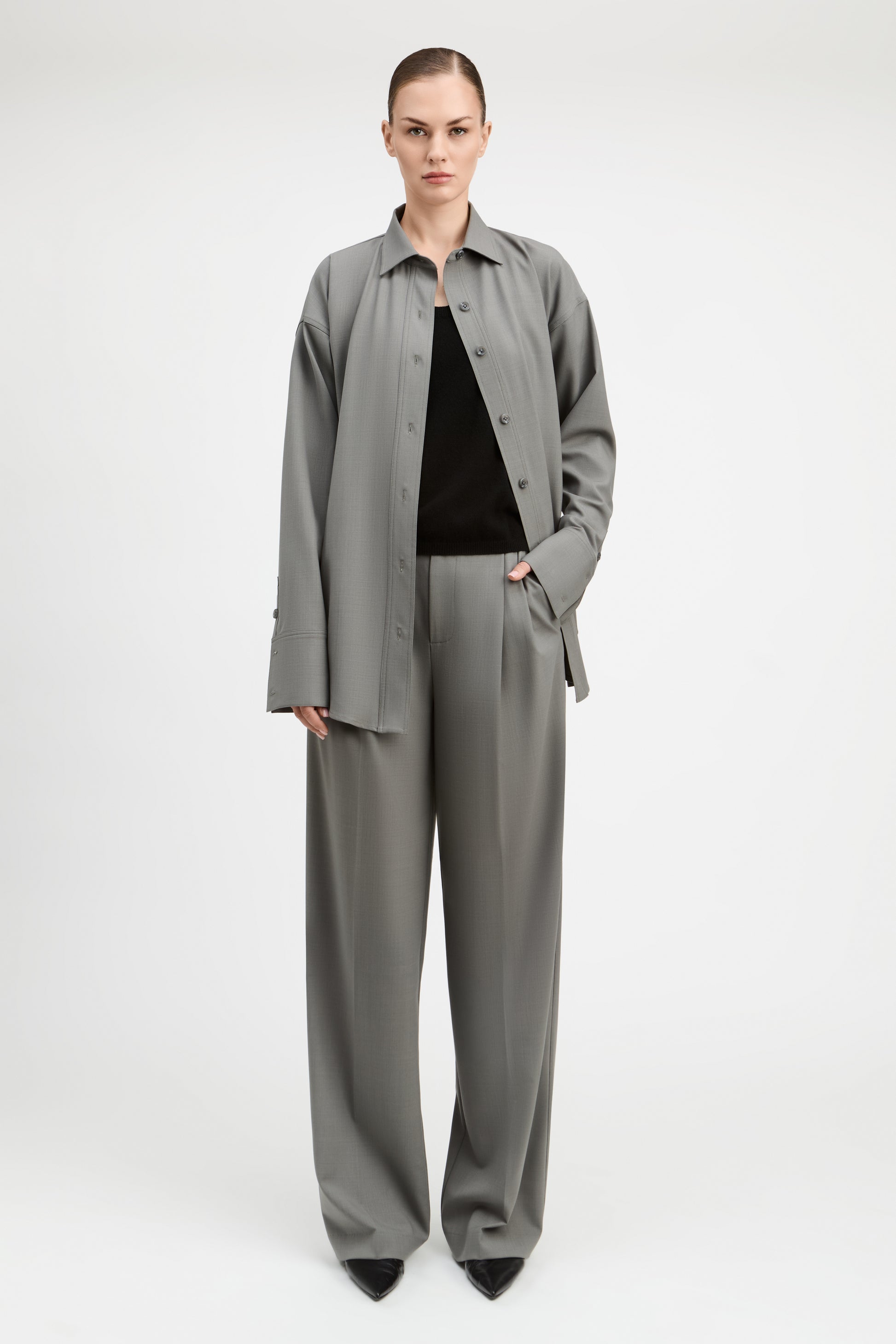 A model stands against a light gray studio background wearing a matching gray two-piece set consisting of a long-sleeve button-front overshirt and wide-leg trousers, styled over a black top and black pointed shoes.