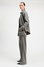 A model is shown from the side wearing a matching gray two-piece suit consisting of a long-sleeve button-up shirt jacket and wide-leg trousers, styled with a black top and black pointed shoes against a plain light gray studio background.