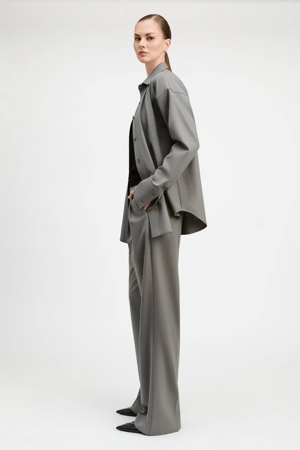 A model is shown from the side wearing a matching gray two-piece suit consisting of a long-sleeve button-up shirt jacket and wide-leg trousers, styled with a black top and black pointed shoes against a plain light gray studio background.