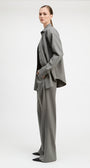 A model is shown from the side wearing a matching gray two-piece suit consisting of a long-sleeve button-up shirt jacket and wide-leg trousers, styled with a black top and black pointed shoes against a plain light gray studio background.