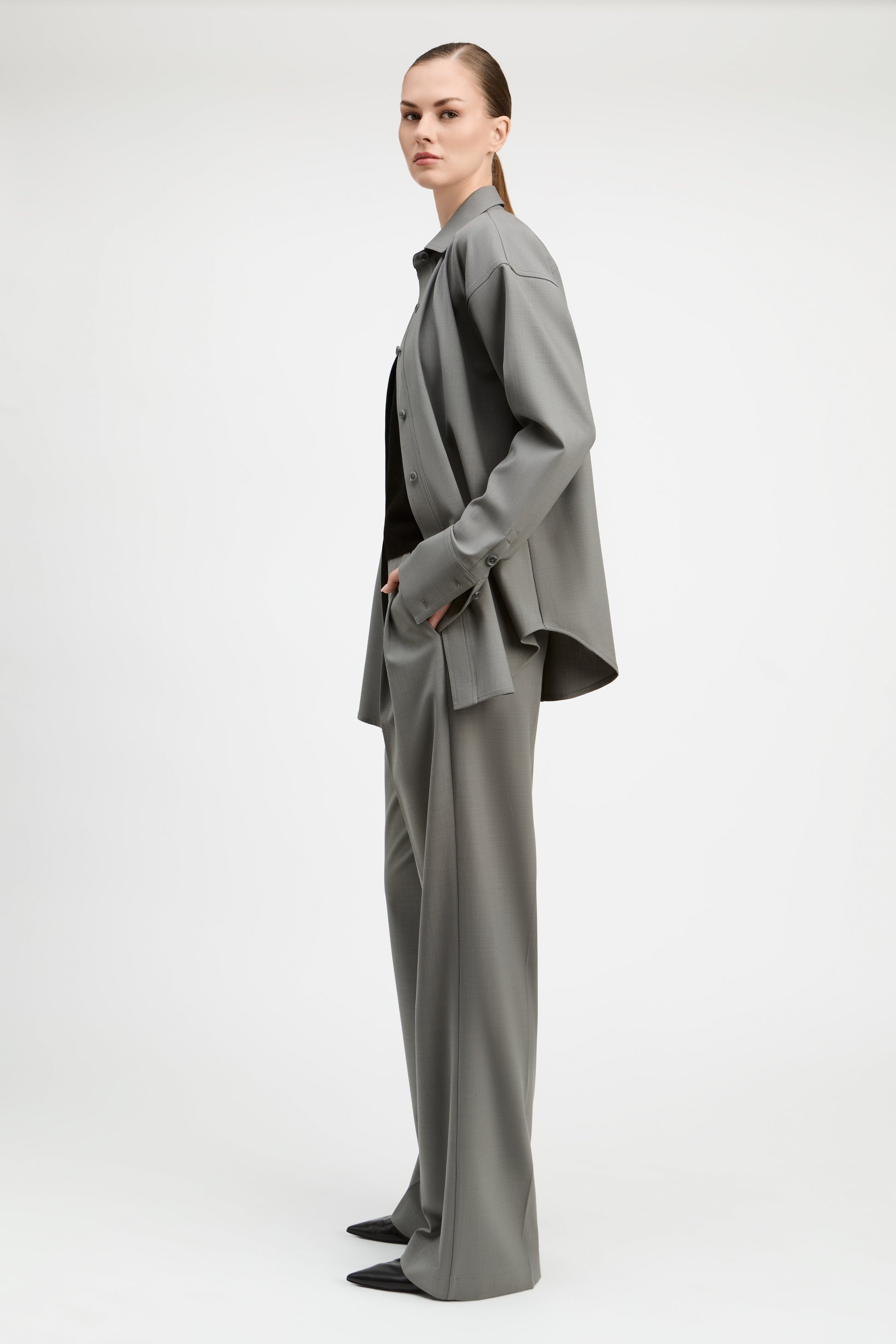 A model is shown from the side wearing a matching gray two-piece suit consisting of a long-sleeve button-up shirt jacket and wide-leg trousers, styled with a black top and black pointed shoes against a plain light gray studio background.