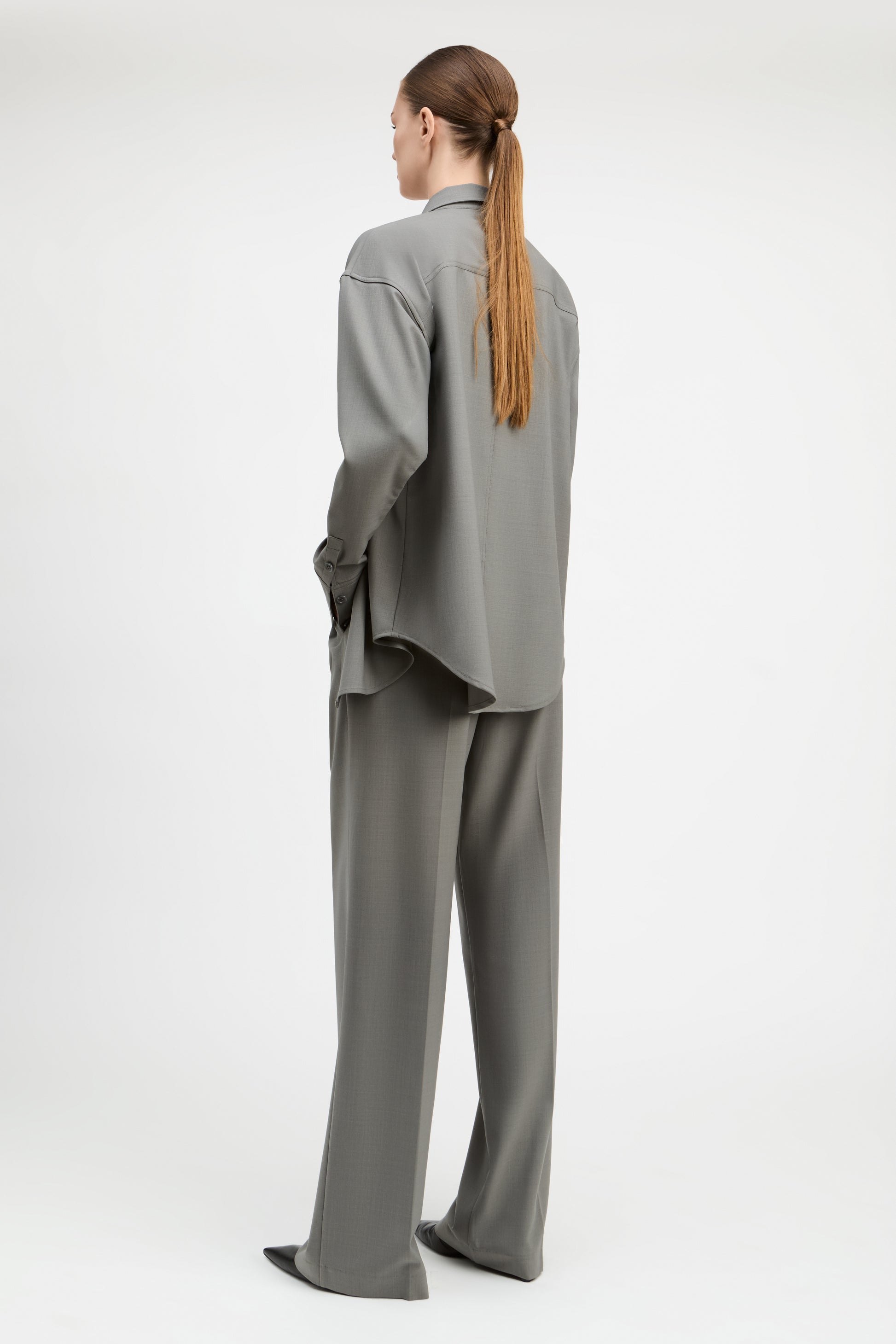 A model shown from the back wearing a matching gray two-piece lounge set consisting of a long-sleeve button-up shirt and wide-leg trousers, photographed against a seamless white studio background.