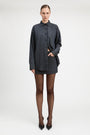 A model stands against a light gray studio background wearing a tailored double-breasted blazer and matching mini skirt in a dark blue and black check pattern, paired with sheer black tights and black pointed heels.