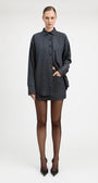 A model stands against a light gray studio background wearing a tailored double-breasted blazer and matching mini skirt in a dark blue and black check pattern, paired with sheer black tights and black pointed heels.