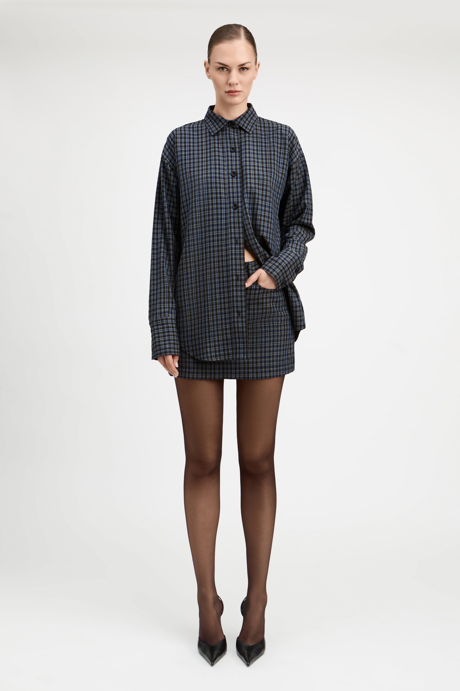 A model stands against a light gray studio background wearing a tailored double-breasted blazer and matching mini skirt in a dark blue and black check pattern, paired with sheer black tights and black pointed heels.