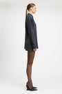 A model stands against a light gray studio background wearing a tailored double-breasted blazer and matching mini skirt in a dark blue and black check pattern, paired with sheer black tights and black pointed heels.