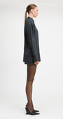 A model stands against a light gray studio background wearing a tailored double-breasted blazer and matching mini skirt in a dark blue and black check pattern, paired with sheer black tights and black pointed heels.