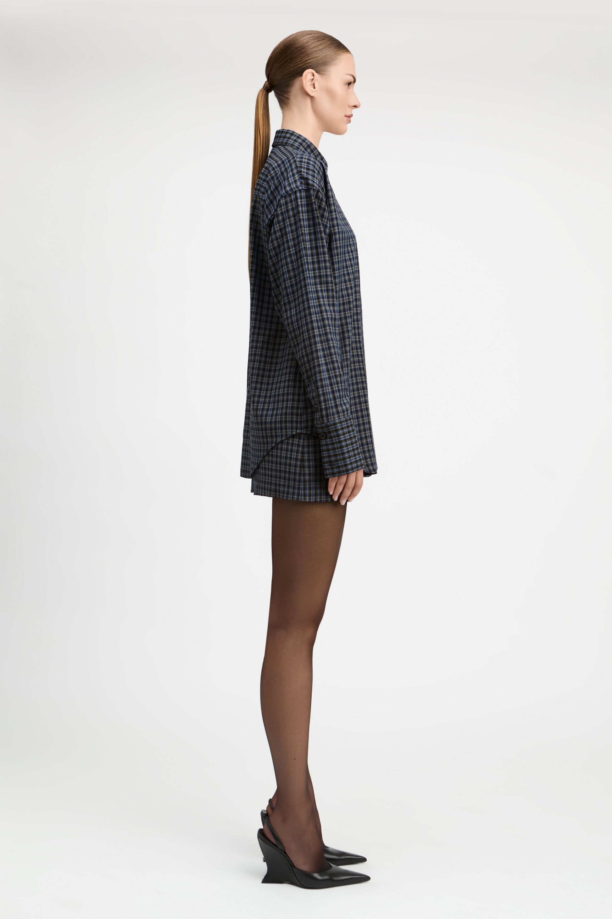 A model stands against a light gray studio background wearing a tailored double-breasted blazer and matching mini skirt in a dark blue and black check pattern, paired with sheer black tights and black pointed heels.