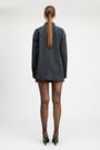 A model stands against a light gray studio background wearing a tailored double-breasted blazer and matching mini skirt in a dark blue and black check pattern, paired with sheer black tights and black pointed heels.