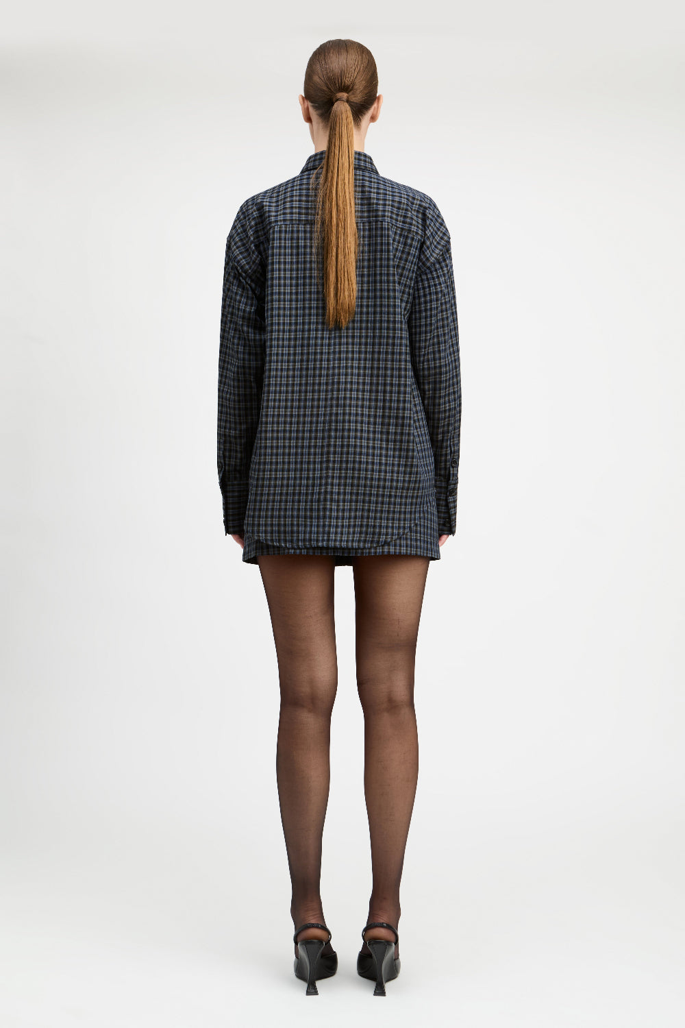 A model stands against a light gray studio background wearing a tailored double-breasted blazer and matching mini skirt in a dark blue and black check pattern, paired with sheer black tights and black pointed heels.