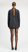 A model stands against a light gray studio background wearing a tailored double-breasted blazer and matching mini skirt in a dark blue and black check pattern, paired with sheer black tights and black pointed heels.