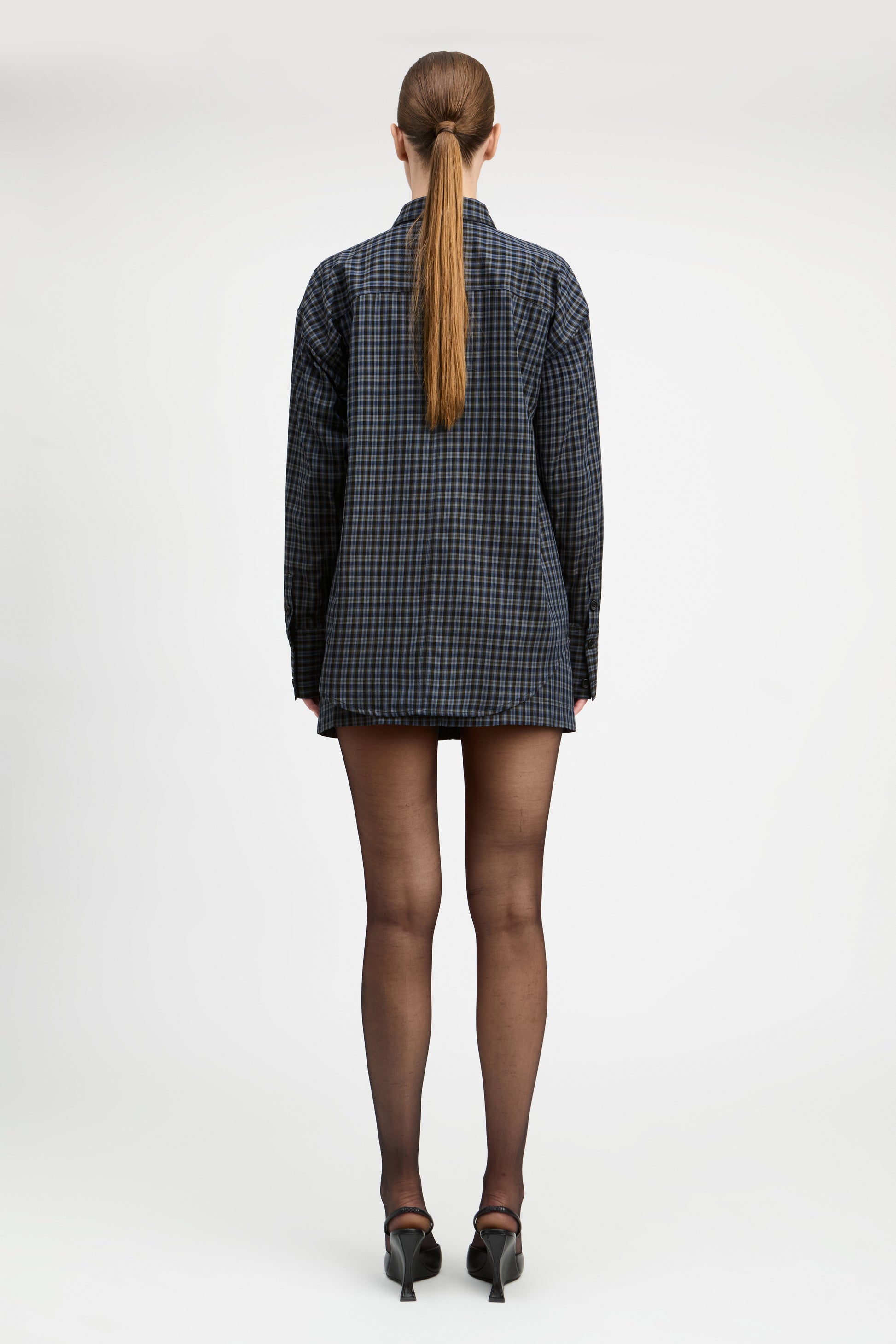 A model stands against a light gray studio background wearing a tailored double-breasted blazer and matching mini skirt in a dark blue and black check pattern, paired with sheer black tights and black pointed heels.