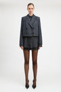 A model stands against a light gray studio background wearing a tailored double-breasted blazer and matching mini skirt in a dark blue and black check pattern, paired with sheer black tights and black pointed heels.