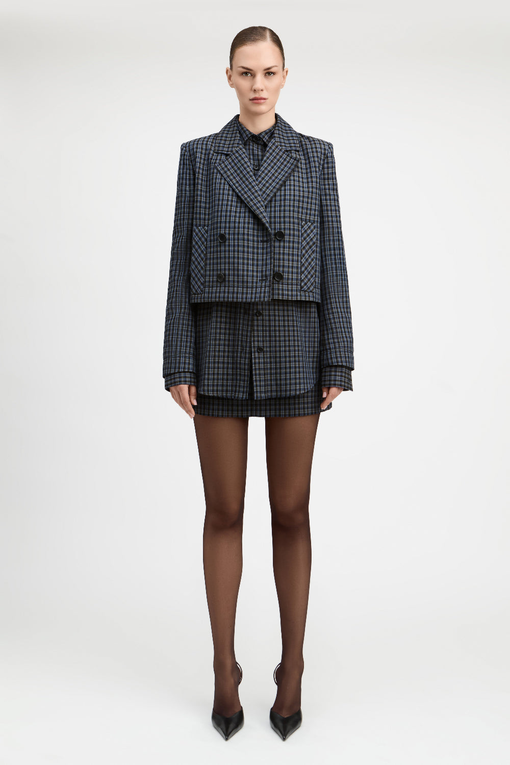 A model stands against a light gray studio background wearing a tailored double-breasted blazer and matching mini skirt in a dark blue and black check pattern, paired with sheer black tights and black pointed heels.