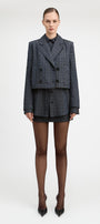 A model stands against a light gray studio background wearing a tailored double-breasted blazer and matching mini skirt in a dark blue and black check pattern, paired with sheer black tights and black pointed heels.