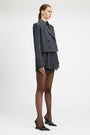 A model stands against a light gray studio background wearing a tailored double-breasted blazer and matching mini skirt in a dark blue and black check pattern, paired with sheer black tights and black pointed heels.