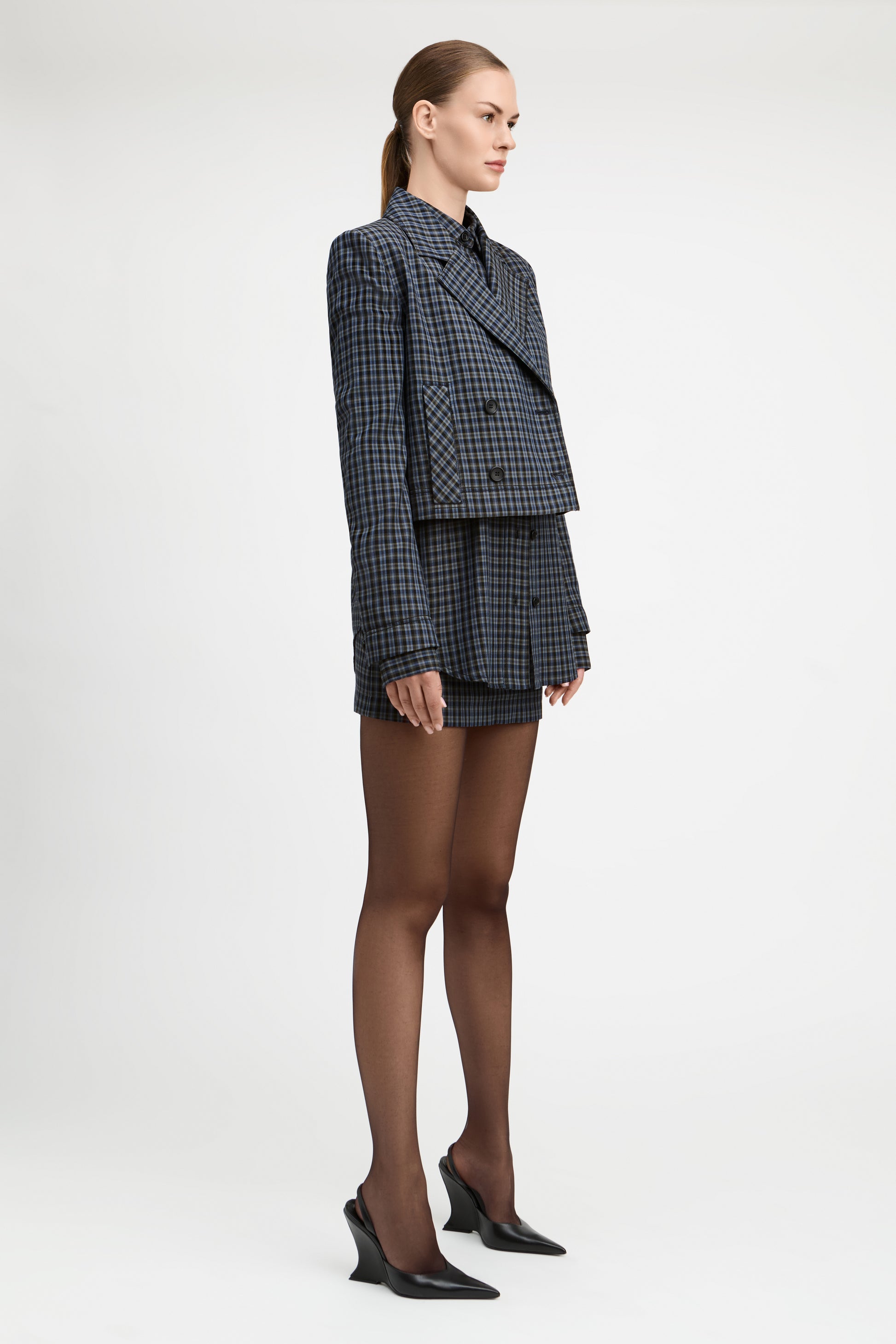 A model stands against a light gray studio background wearing a tailored double-breasted blazer and matching mini skirt in a dark blue and black check pattern, paired with sheer black tights and black pointed heels.