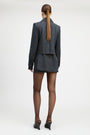 A model stands against a light gray studio background wearing a tailored double-breasted blazer and matching mini skirt in a dark blue and black check pattern, paired with sheer black tights and black pointed heels.