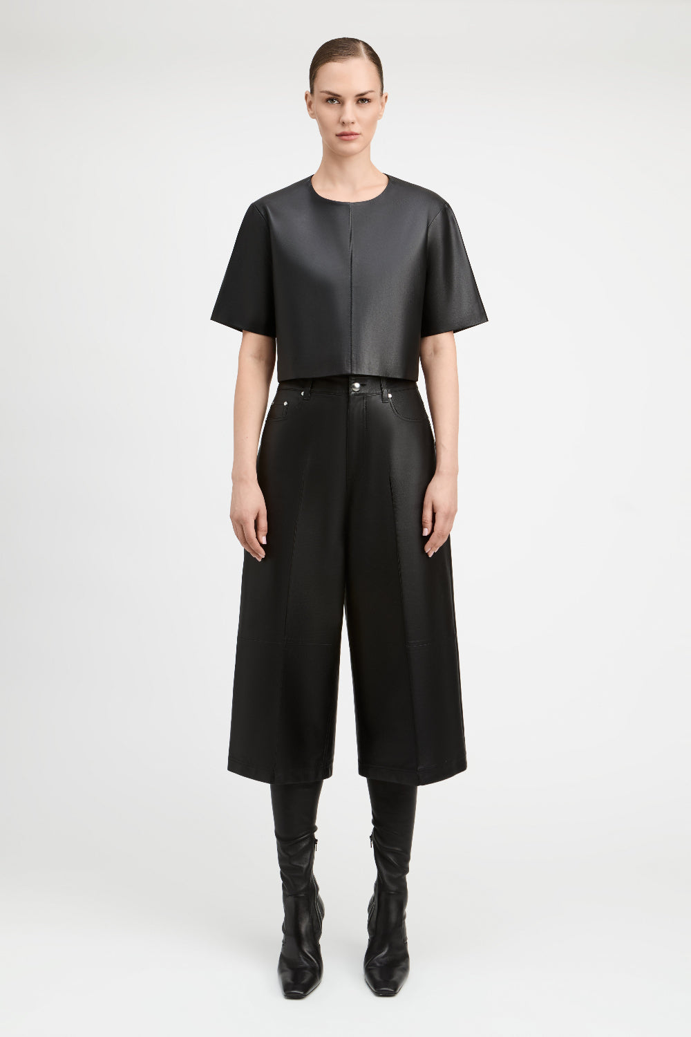 A model stands against a light gray studio background wearing a black short-sleeve cropped top and black wide-leg cropped pants with black knee-high boots.