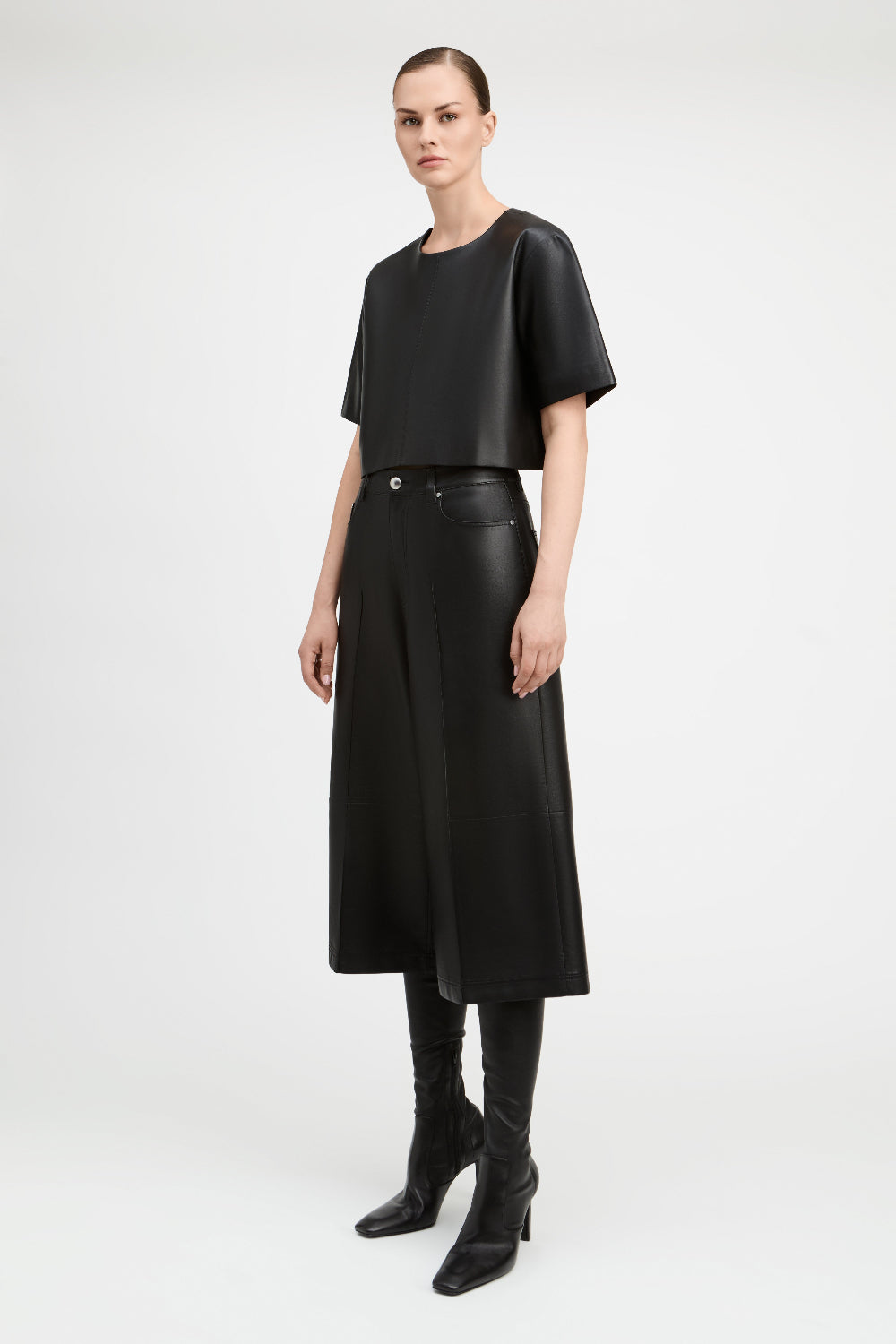 A model stands against a light gray studio background wearing a black short-sleeve cropped top, black high-waisted wide-leg skirt, black skinny boots, and black leather-look belt with a silver buckle.